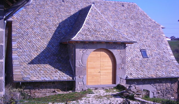 Toiture en lauzes traditionnelles du Cantal réalisée par Auritoit, avec porte cintrée en bois et murs en pierre