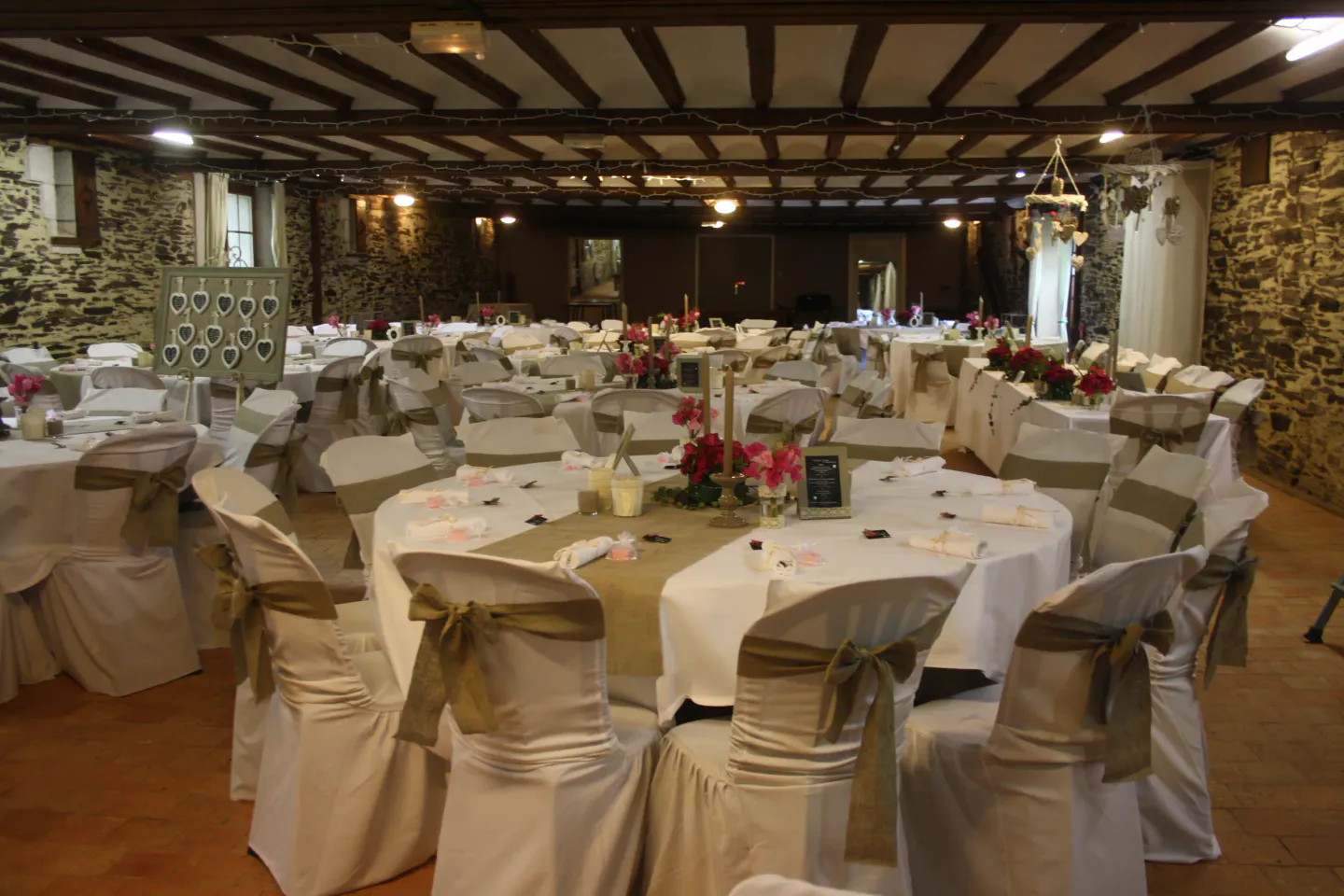 La grande salle de réception des Chais de Rousson : murs de pierre, poutres apparentes et tables dressées en blanc sous la lumière chaude des spots.