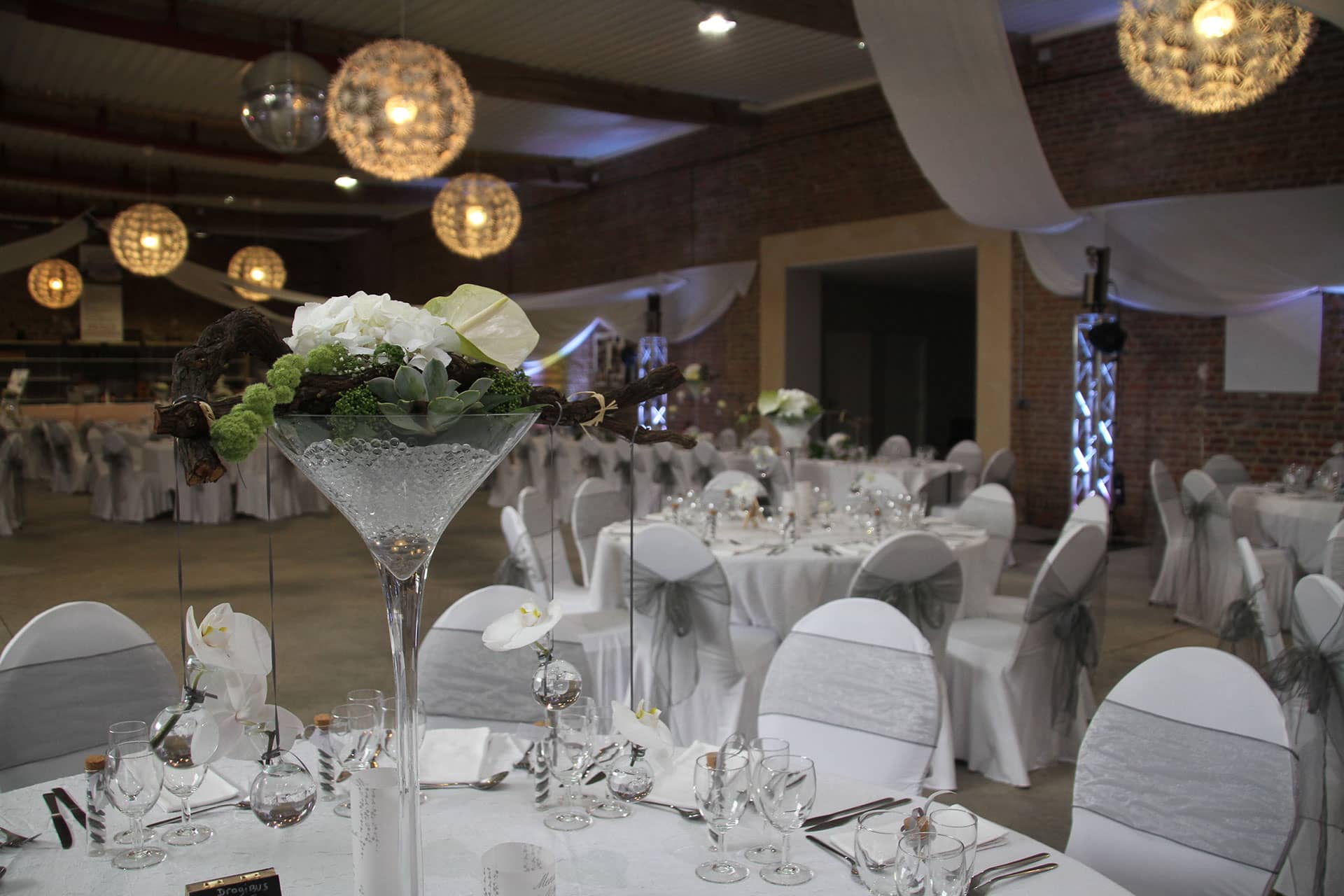La grande salle dressée pour une réception : tables rondes, chaises habillées et compositions florales