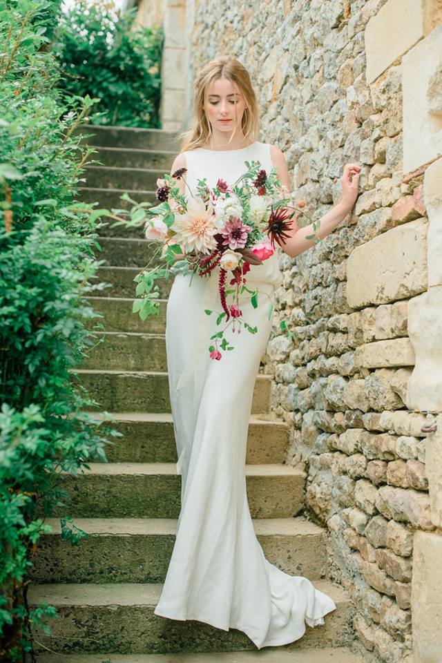 Mariee en robe longue tenant un bouquet, sur un escalier de pierre borde de verdure