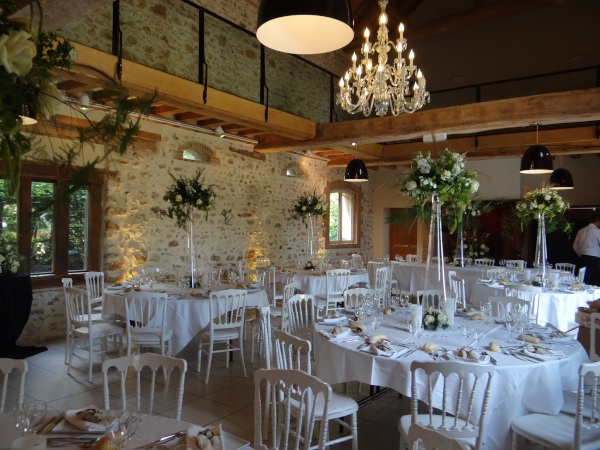 Salle de réception de La Grange des Mollières : pierres et charpente apparentes, plafond cathédrale, lustre et tables de mariage dressées en blanc.