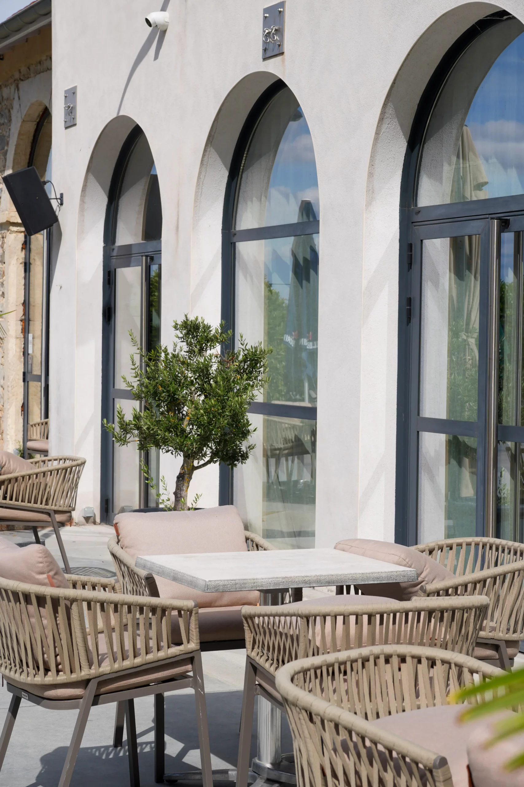 Terrasse extérieure avec grandes arches vitrées, fauteuils en rotin et olivier