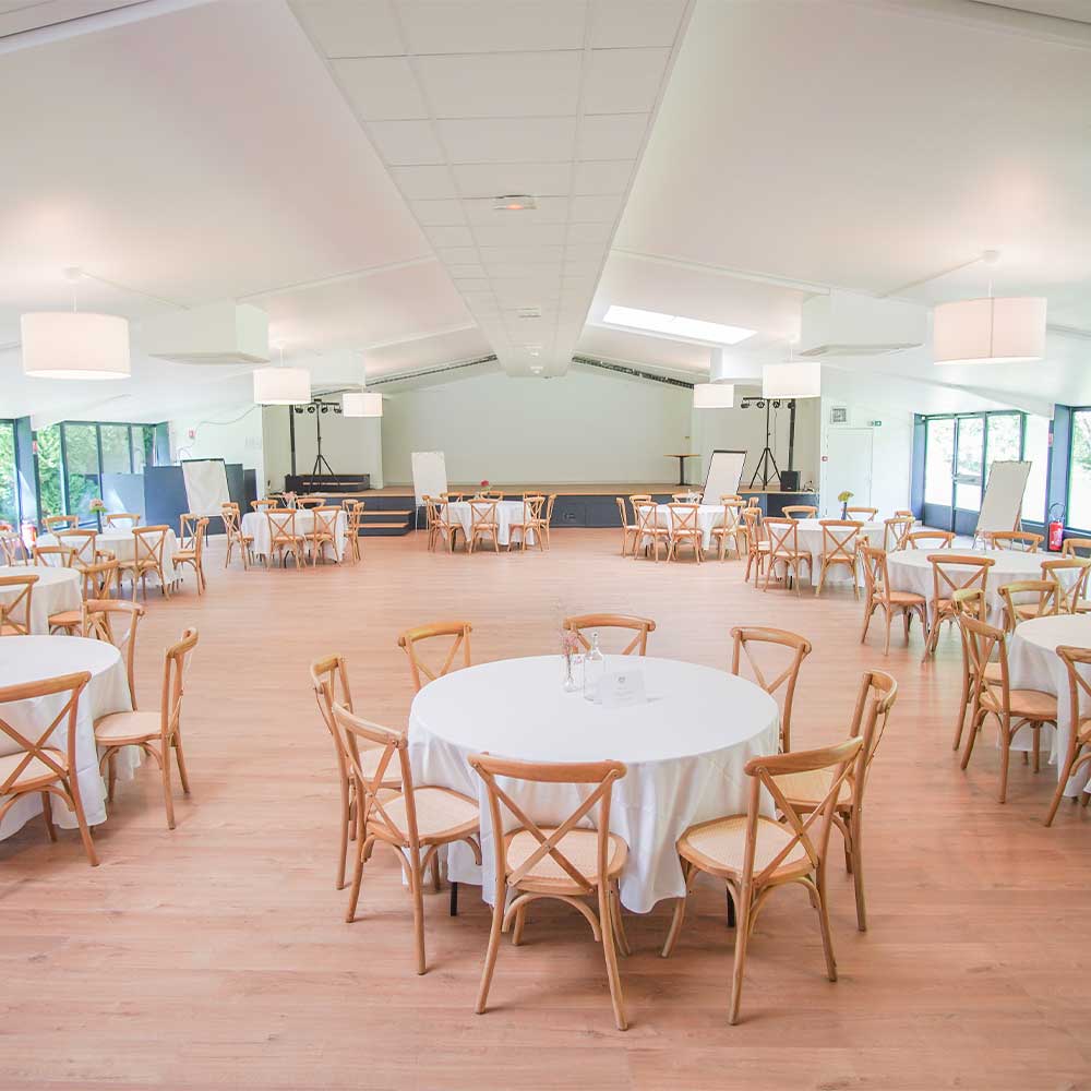 Grande salle de réception lumineuse, charpente claire, tables rondes et chaises bistrot en bois, aux Domaines de Gaston