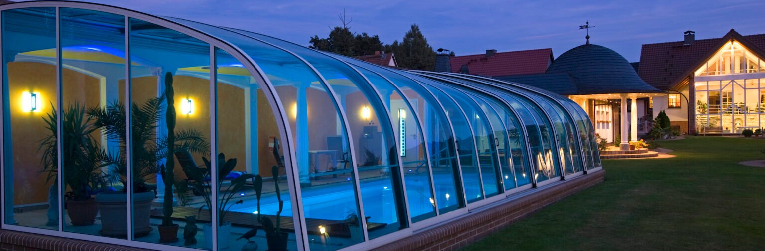 Piscine couverte d'un abri haut au crépuscule, éclairée en bleu, devant une maison de caractère.