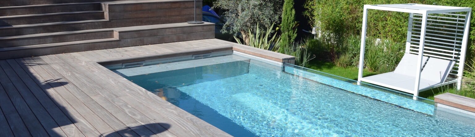 Piscine contemporaine bordée d'une terrasse bois, transat sous pergola dans un jardin paysagé.