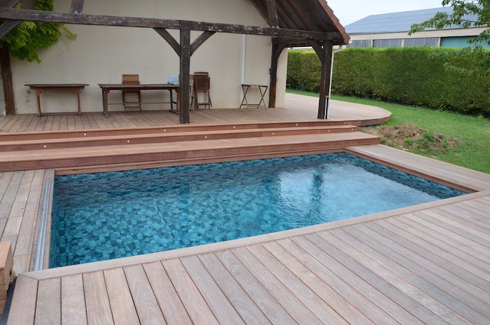 Piscine à mosaïque sombre prolongée par un pool-house couvert et une terrasse en bois.