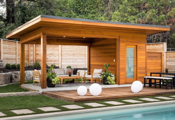 Pool-house en bois éclairé au crépuscule, salon extérieur et boules lumineuses au bord du bassin