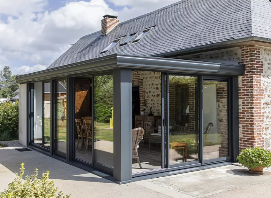 Véranda Soko sur-mesure en aluminium anthracite, baignée de lumière, posée sur une maison de pierre